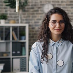 Smiling young woman in stylish office setting, wearing eyeglasses and patterned shirt.